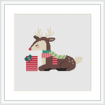 The image features a cross stitch pattern of a stylized seated deer with antlers, wearing a scarf, and sitting beside a wrapped present. The background is plain white, highlighting the colorful design of the deer and the gift.