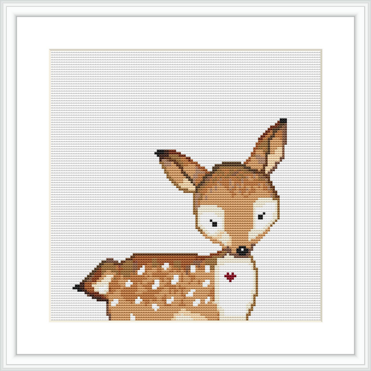 The image depicts a framed cross stitch pattern of a cartoon-like deer with white spots and a red nose. The background is white, and the deer is centered within the frame which is also shown in the image.