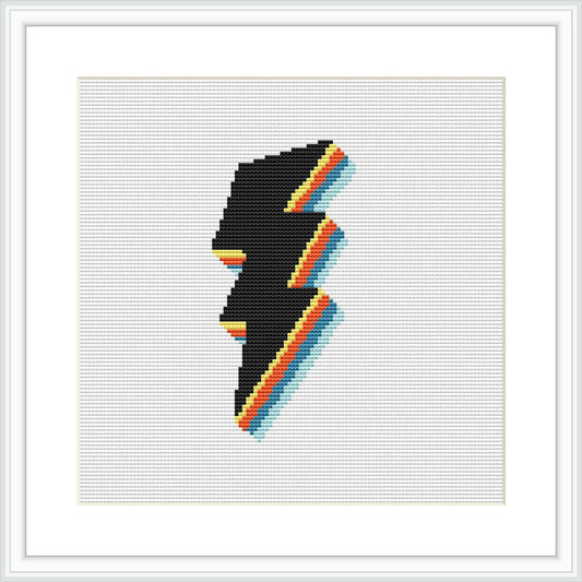 The image displays a cross stitch pattern framed and mounted on a wall. The pattern consists of a stylized silhouette of a face in profile outlined in black and segmented with stripes of bright yellow, orange, and blue against a white background.