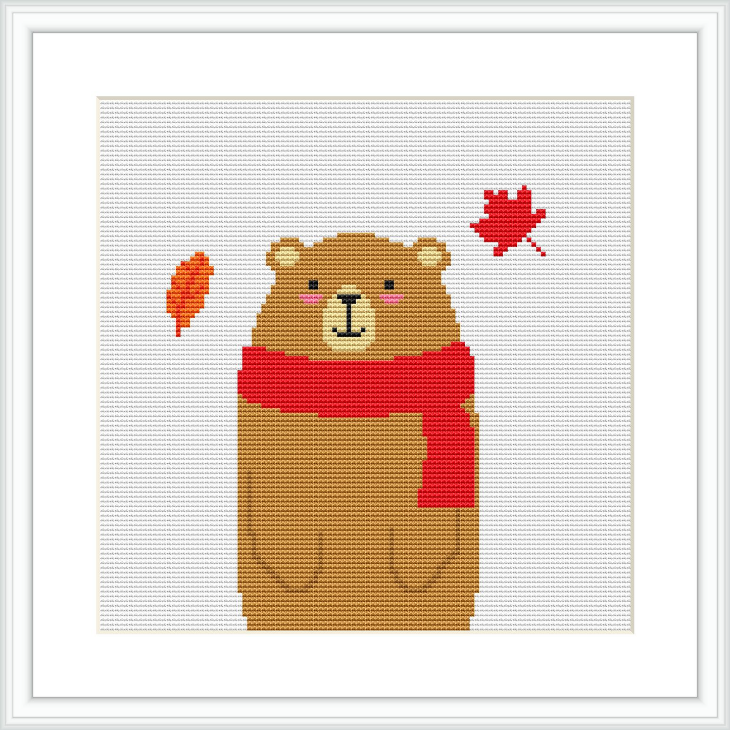 The image depicts a cross stitch pattern of a brown bear with a happy face, wearing a red scarf. There are two autumn leaves, one orange and one red, floating to the bear's right side. The background is white and the design is framed within a simulated picture frame.