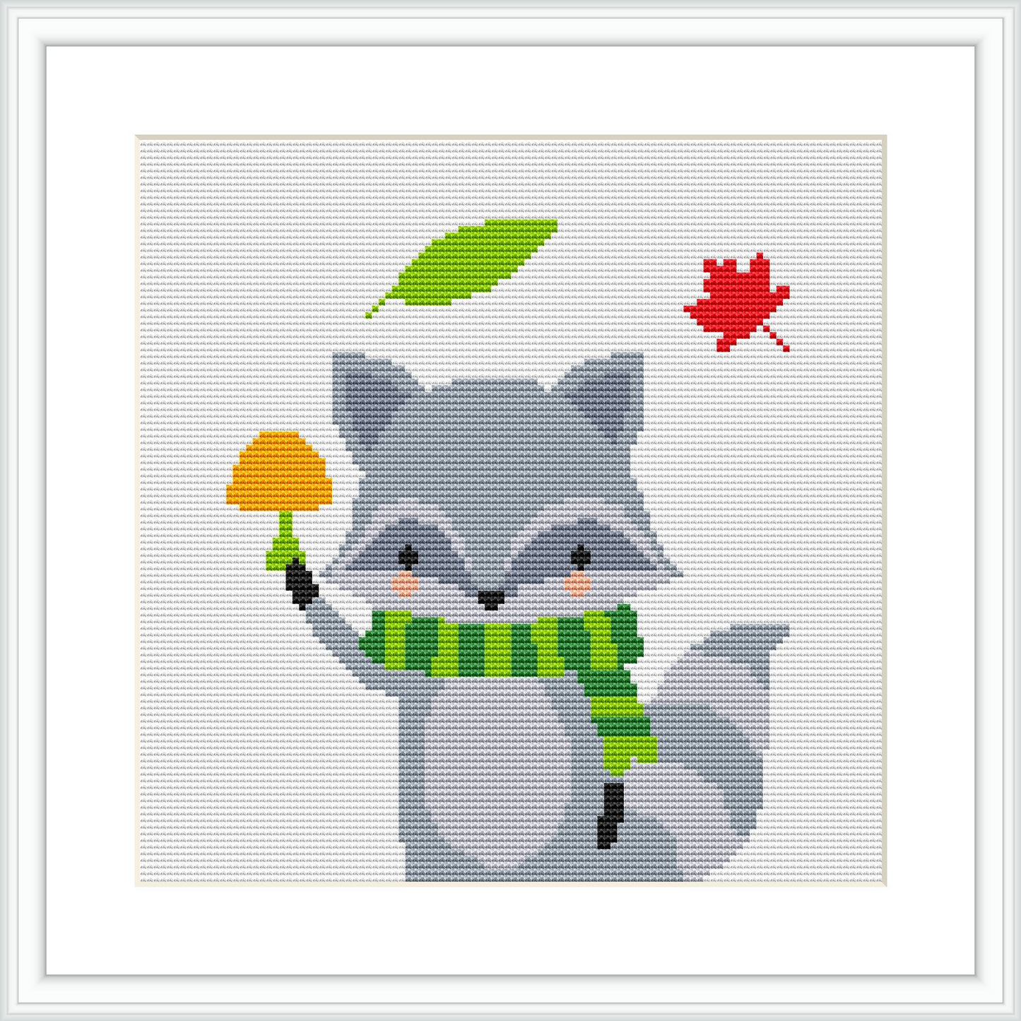 The image shows a cross stitch pattern of a gray raccoon with a green scarf around its neck, holding an orange pumpkin, with a green leaf on its head and a red leaf falling nearby. The raccoon is smiling and appears in a frontal, slightly left-angled pose.