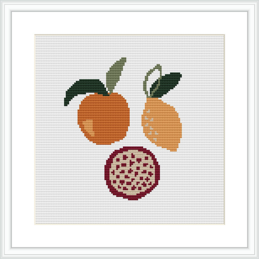 The image presents a framed cross stitch pattern with three fruits displayed vertically; an orange at the top, a lemon in the middle, and a cross-sectioned pomegranate at the bottom set against a white background.