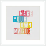 The image shows a framed cross stitch design featuring the phrase 'MAKE YOUR OWN MAGIC' in bold, block letters. Each word is in a different pastel color and staggered for visual interest against a white background.