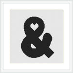 The image depicts a framed cross stitch pattern showcasing the ampersand '&' character with a simple heart shape incorporated into its upper loop. The design is stitched in black on a white canvas and is centered within the frame.