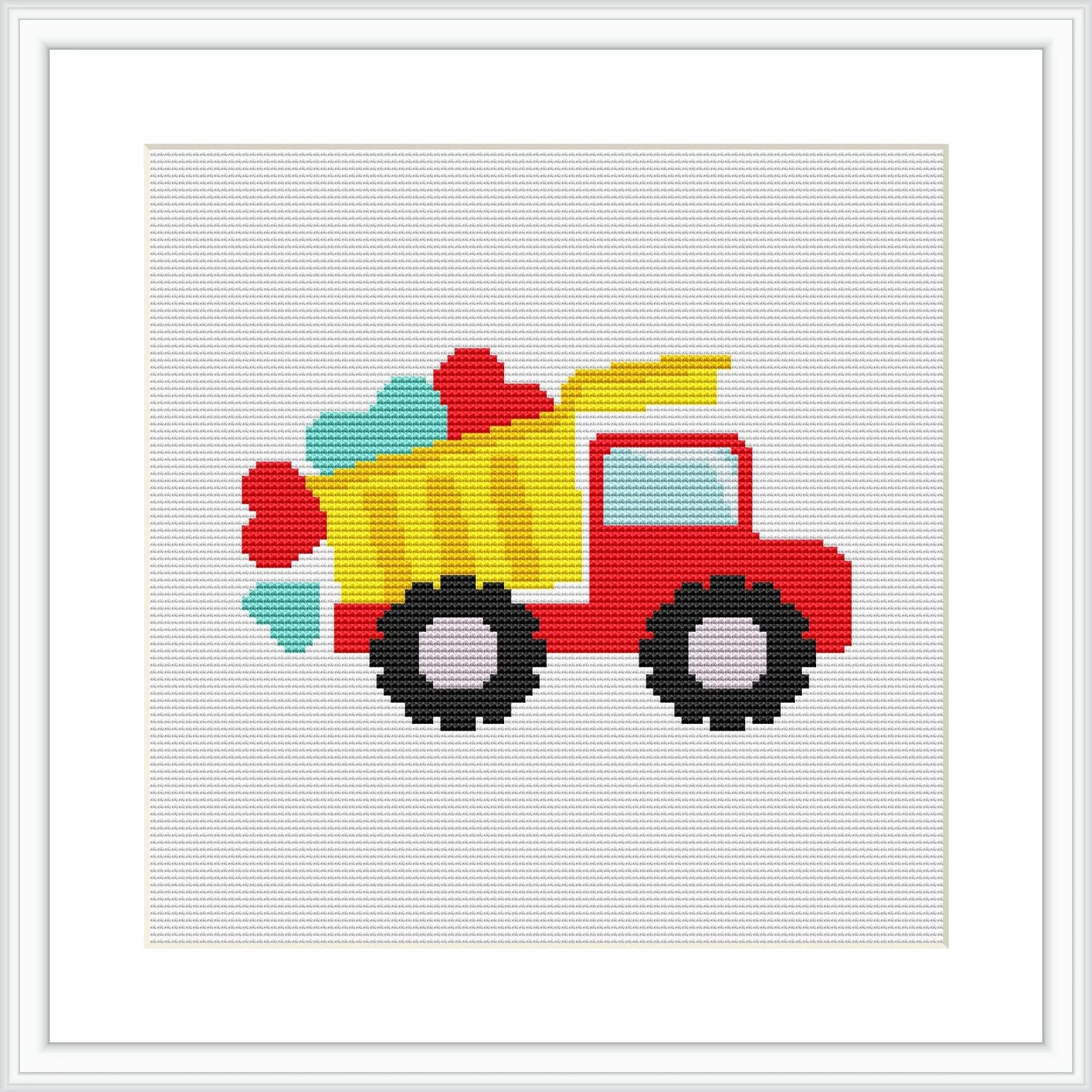 The image features a stylized, colorful cross stitch pattern of a red truck with a yellow bed, carrying multicolored shapes. Black wheels and window, with teal accents, mounted on a white background framed in white.