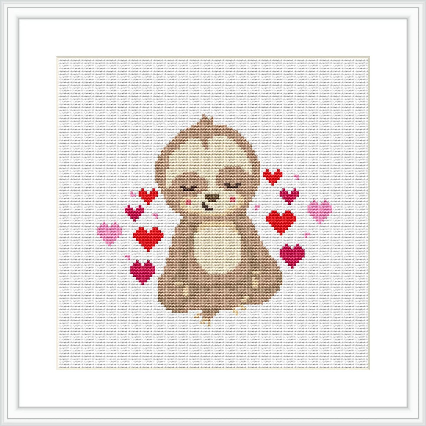 A framed cross stitch piece depicting a brown sloth in a seated meditative pose, with multiple red and pink hearts of various sizes floating around it against a white background.