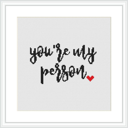 The image features black cross-stitched text that reads 'you're my person' with a small red heart at the bottom right, all centered on a white aida cloth, framed in a simple square frame.