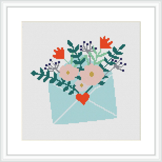 The image depicts a cross stitch pattern featuring an envelope in a soft pastel blue, with a variety of flowers blooming out of its open flap. The flowers include shades of pink, orange, and lavender with green leaves. There is a small red heart in the center of the arrangement, and the envelope appears to be on a plain, lightly shaded background within a square frame.