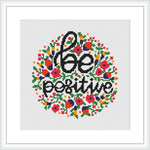 The image displays a cross stitch pattern featuring the phrase 'Be Positive' at the center, surrounded by a circular floral border. Various colors are used to create the flowers and leaves, which are symmetrically arranged around the central text. The pattern is presented on a white background within a framed piece.