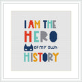 The image features a cross stitch pattern with a phrase that reads 'I AM THE HERO of my own HISTORY' in colorful block letters. Each line of text is in a different color, and there are graphic elements such as a dotted line, a solid circle, and a stylized crown-like shape above the word 'HERO'. The background is white and the design is framed in a simple, white square frame.