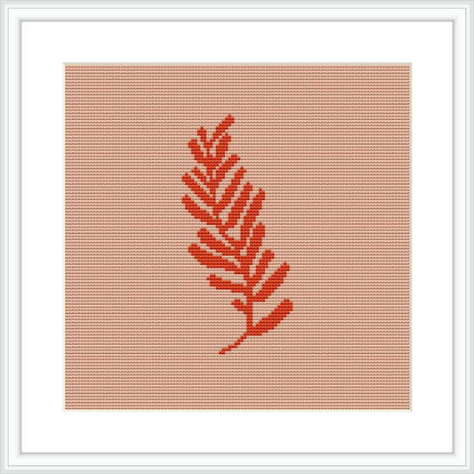 The image presents a single, coral-colored fern leaf centered on an even-weave fabric. The design is framed within a white square border, suggesting it is likely a cross stitch piece ready for display.