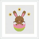 The image shows a pixel art-style cross stitch pattern of a brown Easter bunny with pink inner ears and cheeks, popping out of a decorated pink and green egg. The bunny is centered on a white canvas, with yellow and brown flowers dotted around it.