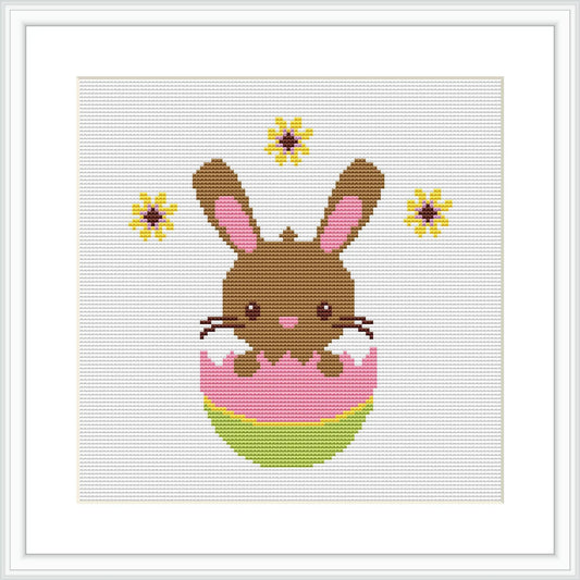 The image shows a pixel art-style cross stitch pattern of a brown Easter bunny with pink inner ears and cheeks, popping out of a decorated pink and green egg. The bunny is centered on a white canvas, with yellow and brown flowers dotted around it.