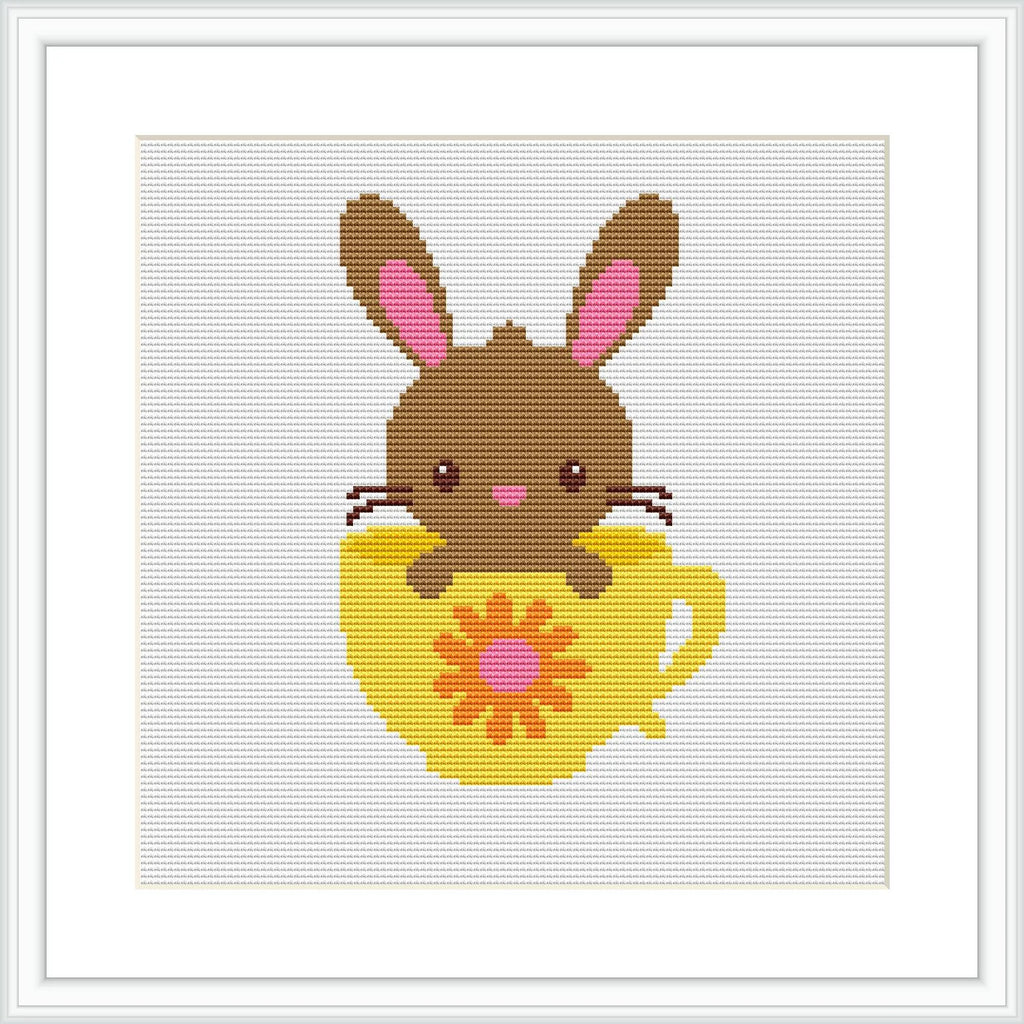 The image shows a cross-stitch pattern depicting a brown bunny with pink inner ears and blushing cheeks sitting inside a yellow egg-shaped cup with a red floral design on it. The background is white, and the pattern is framed with a simple square frame.
