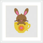 The image shows a cross-stitch pattern depicting a brown bunny with pink inner ears and blushing cheeks sitting inside a yellow egg-shaped cup with a red floral design on it. The background is white, and the pattern is framed with a simple square frame.