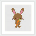 The image shows a framed cross stitch pattern of a cute brown bunny with pink inner ears standing upright. The bunny is holding a carrot with green leaves in its front paws. The background is white, and the stitching is visible.