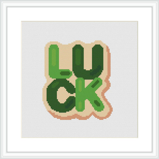 The image features a cross stitch pattern with the word 'LUCK' in green and peach colors, block lettering style, and a white background, all framed in a simple white frame.