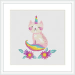 The image showcases a cross stitch pattern of a pink caticorn with a multicolored tail and horn, seated in the center. It is surrounded by flowers with purple petals and yellow centers, set against a plain white background.