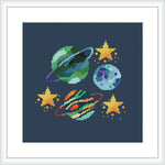 The image depicts a cross stitch pattern framed in white, showcasing a dark blue background with stylized representations of planets in turquoise, orange, and purple, along with yellow stars. The elements are arranged in a scattered, playful manner across the canvas.