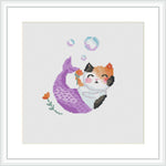 The image depicts a framed cross stitch design of a cat with a mermaid tail. The cat appears to be in the water with bubbles surrounding it, against a white background within a squared frame.