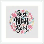 The image displays a cross stitch pattern featuring the phrase 'Best Mom Ever' in stylish, bold letters at the center. Surrounding the text is a decorative wreath made up of flowers and leaves in various colors. The pattern is framed within a white border and presented as if on a wall.