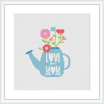 The image features a cross stitch pattern of a blue watering can with the phrase 'Love Grows Here'. Within the can, pink and red flowers are blossoming out, presented on a white background, encased in a simple square frame.