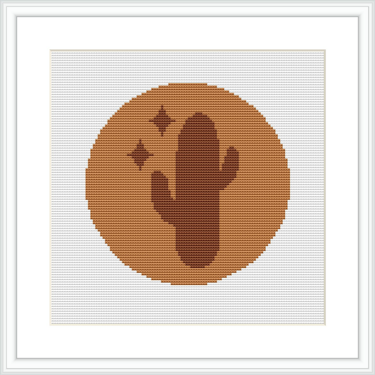 The image displays a framed cross stitch pattern featuring a simple cactus silhouette centered within a circular frame, set against a cream-colored background. The pattern is comprised of small, uniform stitches creating the illustration.