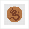 The image features a framed cross stitch pattern with a central monogram symbol resembling a stylized letter 'G' set within a circular frame. The color palette consists of brown tones, creating a warm and inviting look.