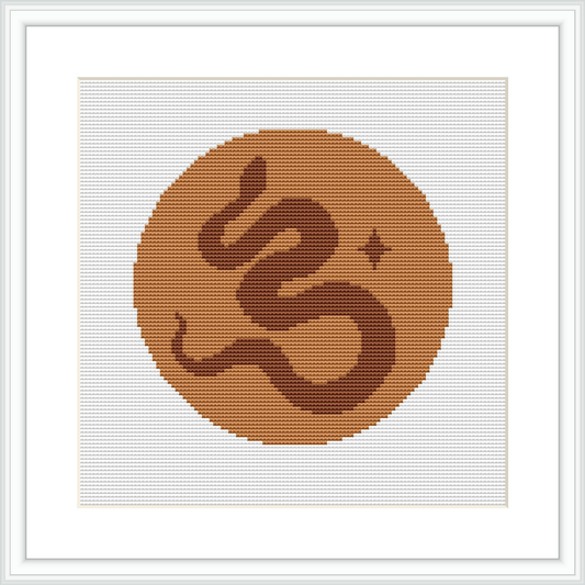 The image features a framed cross stitch pattern with a central monogram symbol resembling a stylized letter 'G' set within a circular frame. The color palette consists of brown tones, creating a warm and inviting look.