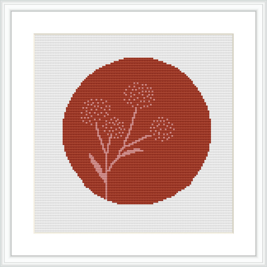 The image shows a cross stitch pattern design of a circular terracotta-colored background with a simple white floral motif at the center, framed by a white border and mounted in a square frame.