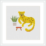 The image shows a cross stitch pattern of a yellow and black spotted leopard sitting next to a green potted plant with pink details on a stool. The background is white, emphasizing the pattern's colors.