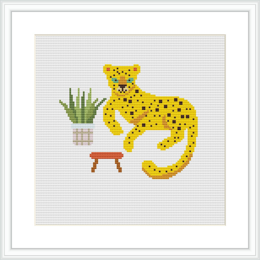 The image shows a cross stitch pattern of a yellow and black spotted leopard sitting next to a green potted plant with pink details on a stool. The background is white, emphasizing the pattern's colors.