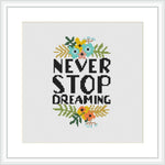 The image depicts a cross stitch pattern framed in white. It features the phrase 'NEVER STOP DREAMING' in capital letters, surrounded by a collection of stylized flowers and leaves at the top and bottom.