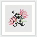 The image shows a framed cross stitch design featuring pink flowers and green leaves, with the phrase 'You make me smile' incorporated into the pattern, all against a white background.