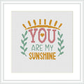 The image displays a framed cross stitch pattern with the phrase 'YOU ARE MY SUNSHINE' in bold, capital letters, accompanied by sun rays at the top and green laurels at the bottom on a white canvas.