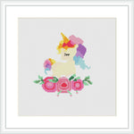 The image depicts a framed cross stitch pattern featuring a unicorn with a rainbow mane and tail, framed by flowers at the bottom.