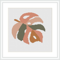 The image shows a cross stitch pattern featuring an abstract botanical shape resembling a leaf in peach and olive tones, framed in a simple white frame against a plain white background.