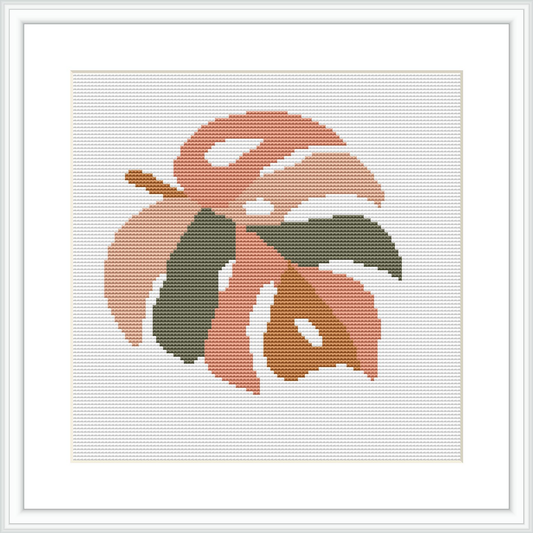 The image shows a cross stitch pattern featuring an abstract botanical shape resembling a leaf in peach and olive tones, framed in a simple white frame against a plain white background.