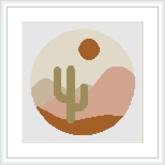 A framed cross stitch pattern showing a stylized desert scene with a cactus, rolling hills and a setting sun within a circle, all rendered in a pixel-like geometric style.