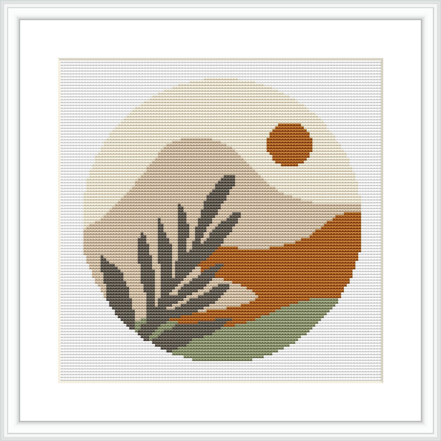 The image depicts a framed cross stitch pattern featuring a circular design with a stylized desert landscape. Elements include a setting sun, mountains, and plants.