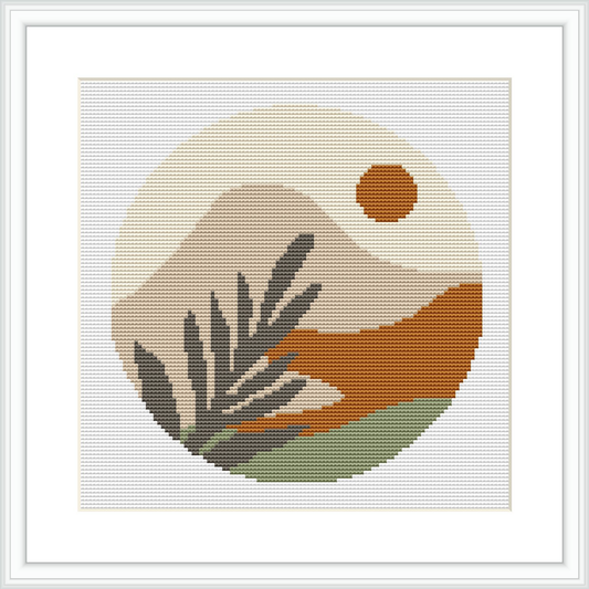 The image depicts a framed cross stitch pattern featuring a circular design with a stylized desert landscape. Elements include a setting sun, mountains, and plants.