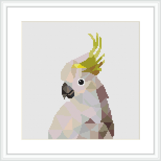 The image shows a framed cross stitch pattern featuring a white cockatoo with a green and yellow crest, perched gracefully. The pattern appears to be displayed on a white surface.