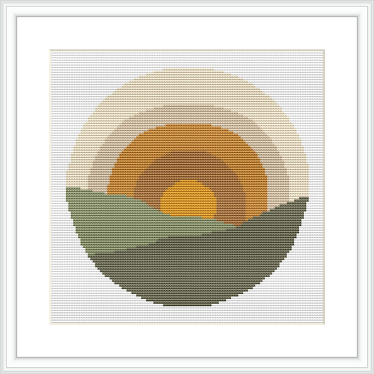 The image shows a cross stitch design featuring a stylized, simplistic sunrise with a yellow center, an orange ring around it, and a larger beige ring enclosing the orange. The bottom half depicts two green hills in a darker tone in front and a lighter tone in the back under a circular backdrop.
