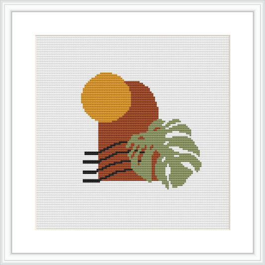 The image shows a framed cross stitch pattern with a centered round element representing the sun in a golden yellow, semi-circular shapes in rust orange below the sun suggesting a landscape, and a green monstera leaf with brown accents to the bottom right, against a plain white background.