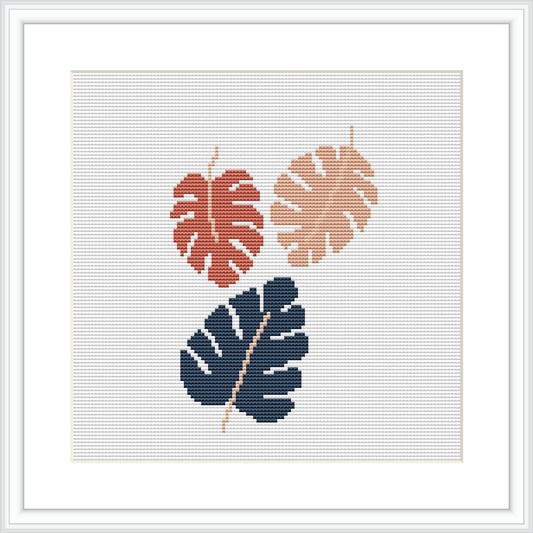 The image shows a cross stitch pattern featuring two leaves—one in burnt orange and the other in muted blue—crossing over each other against an ivory background, framed with a simple white border.