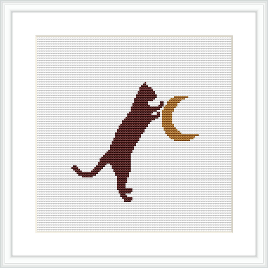 The image features a cross stitch pattern of a brown cat standing on its hind legs, left paw extended upwards, touching a crescent moon. The background is a plain white fabric, framed by a simple white frame, suggesting a wall art decor.