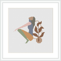 This image showcases a framed cross stitch pattern. It features an abstract silhouette of a woman with an earthy-toned leaf motif. The figure is centered within the square frame, set against a plain white background.