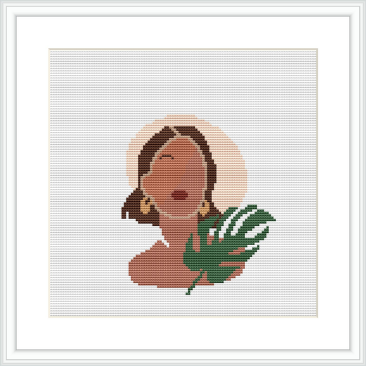 The image shows a framed cross stitch pattern. The pattern depicts a woman's profile with closed eyes and a serene expression. She appears to be wearing a large, round earring and there is a stylized tropical leaf beside her. The background is plain, allowing the figure to stand out.