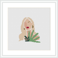 The image depicts a minimalist style cross stitch pattern of a woman with flowing hair and a red lip detail, positioned beside green foliage. The design is displayed within a white frame, placed against a white background.