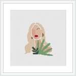 The image depicts a minimalist style cross stitch pattern of a woman with flowing hair and a red lip detail, positioned beside green foliage. The design is displayed within a white frame, placed against a white background.